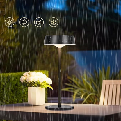Black outdoor lamp with rain simulation on a table with flowers and chair in the background