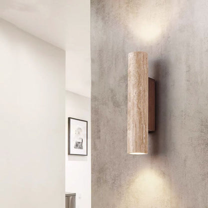 Wall-mounted cylindrical light fixture on a textured wall with a mirror and framed picture in the background.